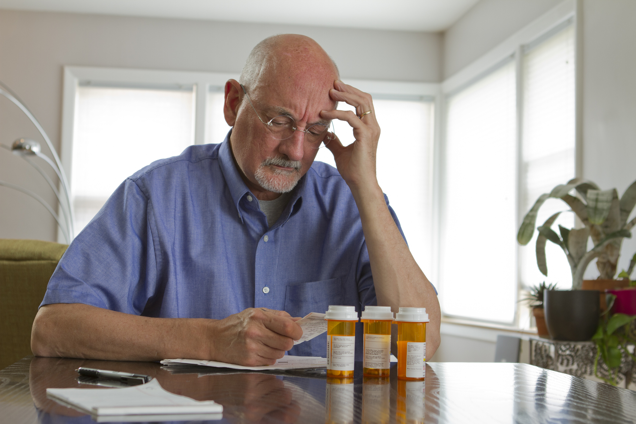 An older white male with prescription drug bottles on his table holds a bill and looks worried.