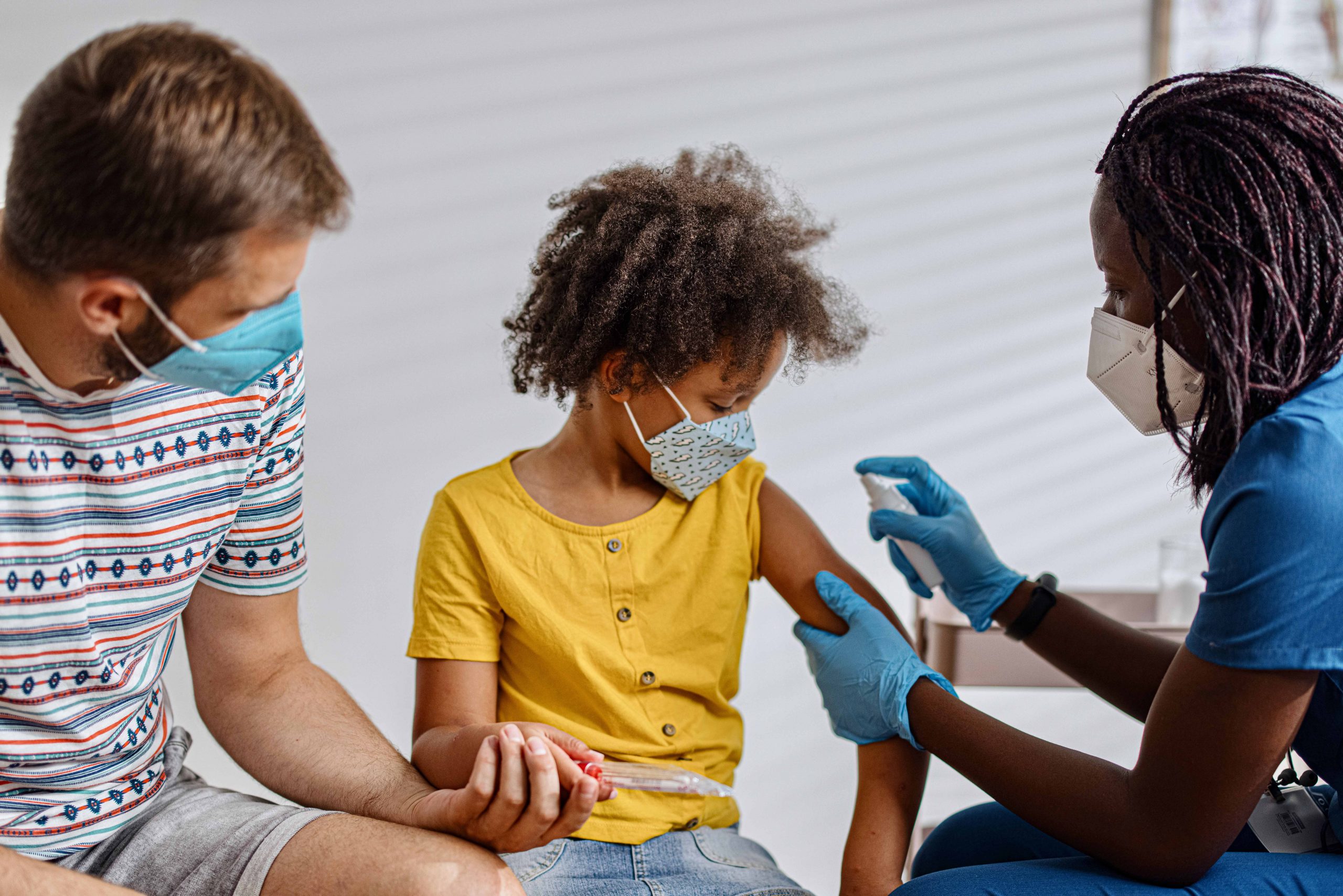 Child with father and female health care professional prepping to provide COVID-19 vaccination.