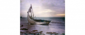 A small, worn looking sailboat abandoned on a beach laying on its side.